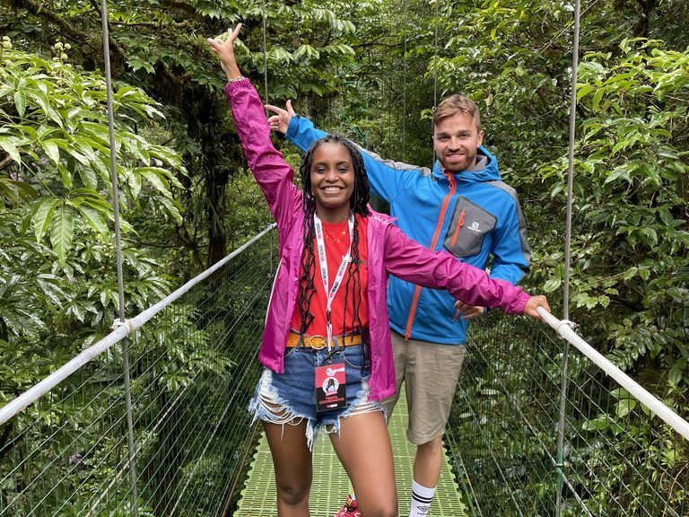 Two smiling travelers posing on a suspension bridge in the lush green rainforest canopy.