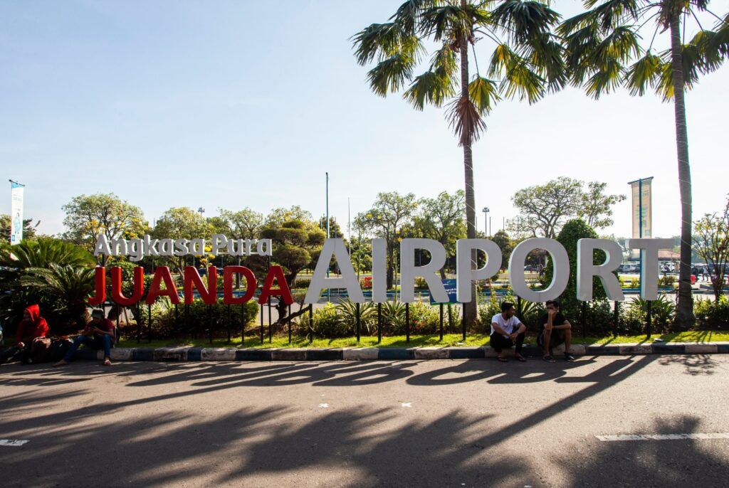 A large outdoor sign for Juanda International Airport surrounded by palm trees and greenery on a sunny day.