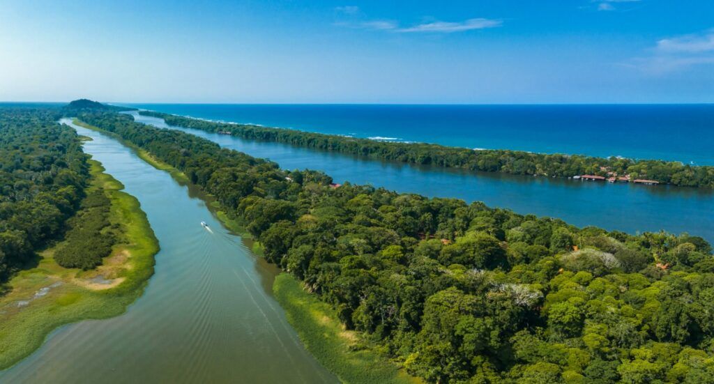 Aerial view of a wide river or canal running parallel to the ocean, separated by a strip of lush green forest.