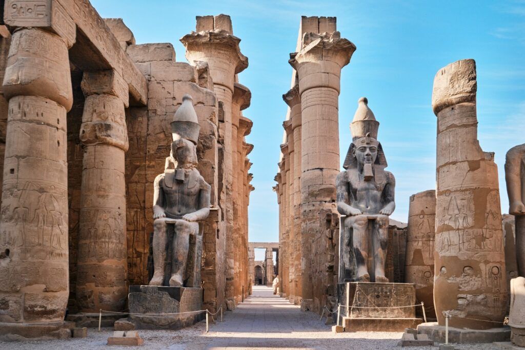 Two massive seated Pharaoh statues standing between large columns in the Amenhotep III Colonnade at the Luxor Temple, Egypt.
