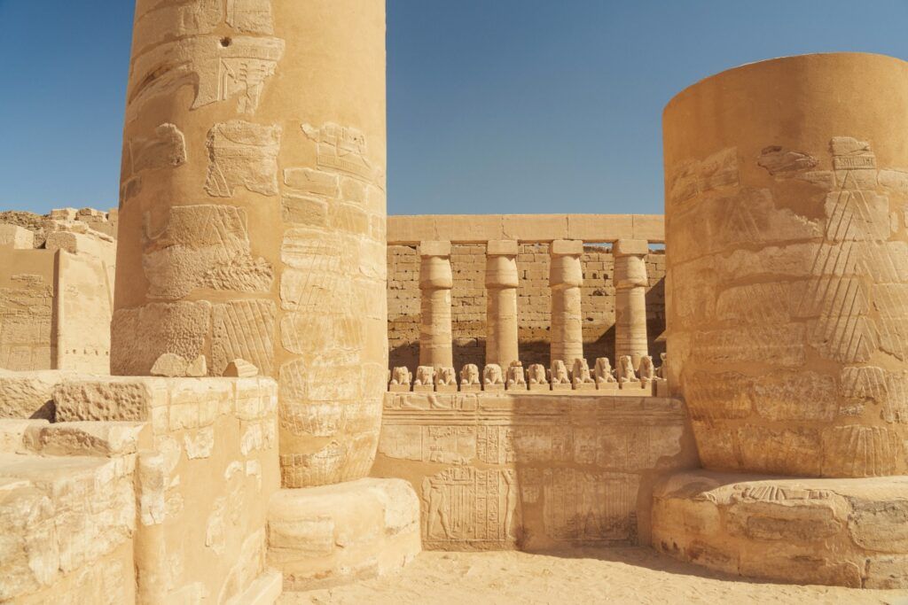 Massive ancient columns with detailed carvings framing a row of smaller columns and stone-carved sphinxes at the Karnak Temple complex.