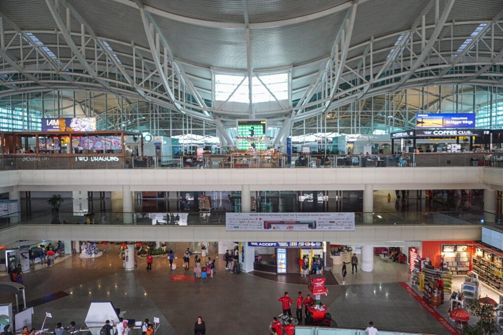 The multi-level interior of the international terminal at Denpasar Airport, featuring a high vaulted ceiling with white structural beams, shops, and passenger lounges.