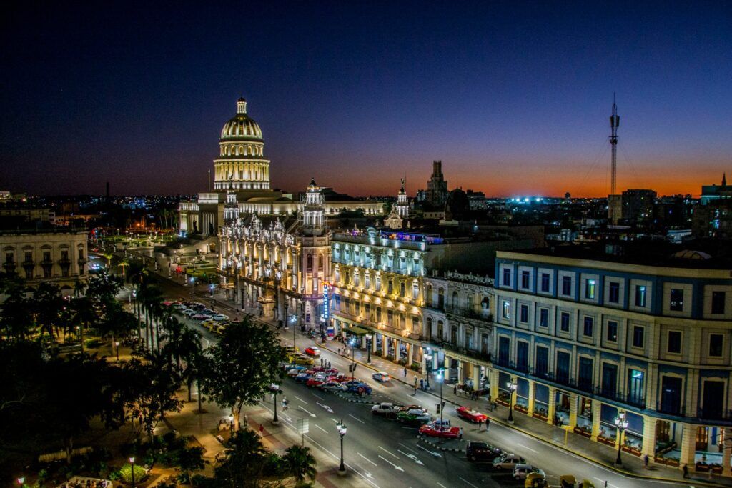 Eine nächtliche Aufnahme aus großer Höhe einer breiten Straße in Havanna mit dem beleuchteten Nationalkapitol und kunstvollen historischen Hotels unter einem tiefblauen Abendhimmel.