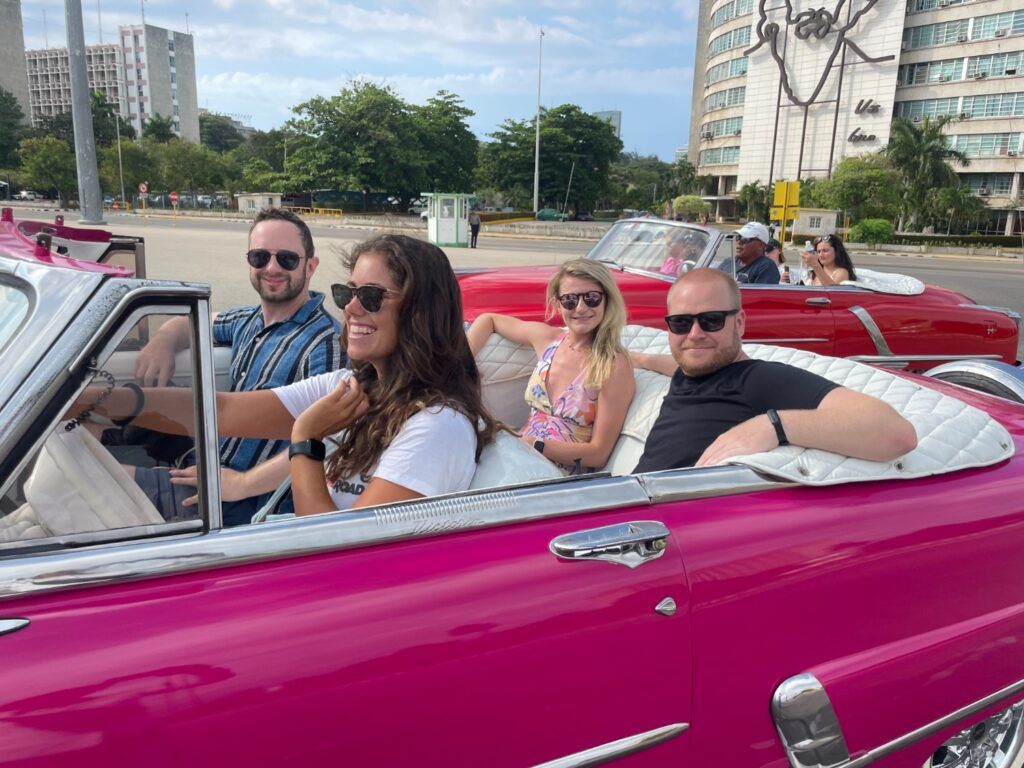 Four happy travelers smiling and enjoying a ride in a bright pink vintage convertible on a city street in Havana, Cuba, with other classic cars in the background.