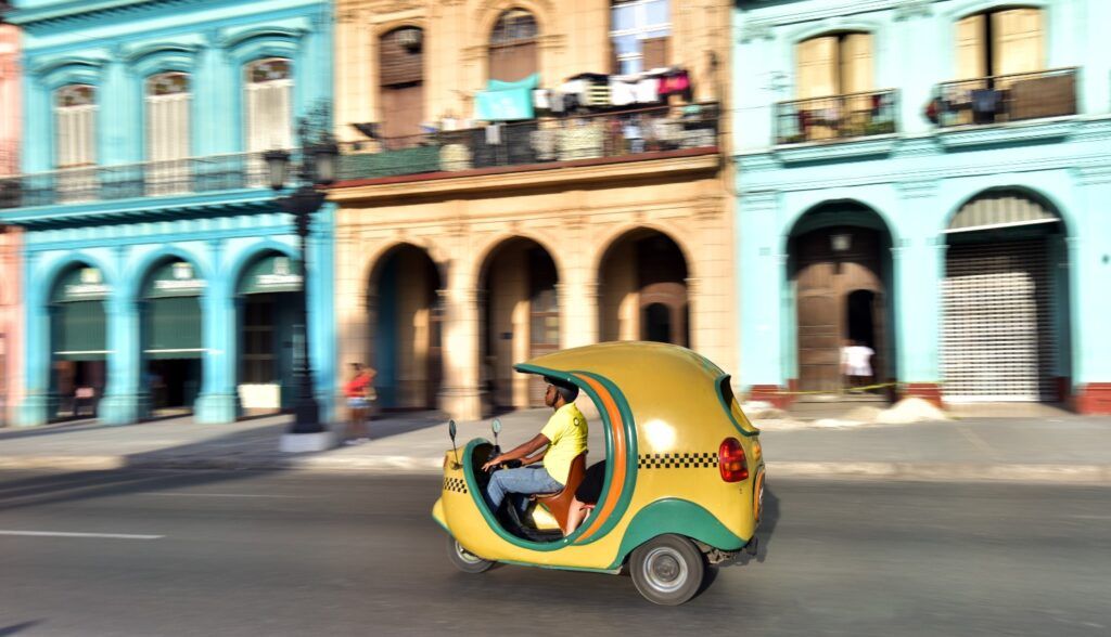 A yellow Coco Taxi driving past colorful colonial buildings on a street in Havana.