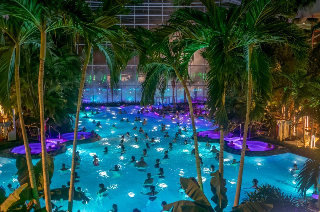 A large indoor thermal pool at Therme Bucharest filled with people, illuminated with blue and purple lights and surrounded by tall palm trees at night.