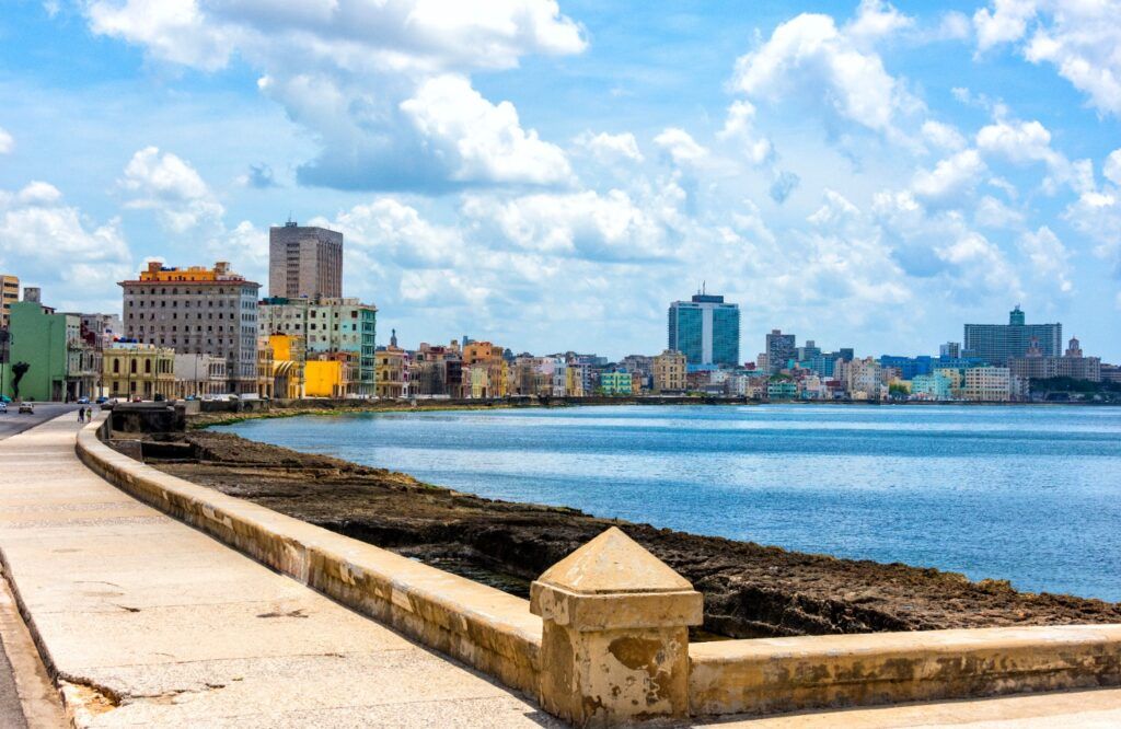 Ein weitläufiger Blick auf die Uferpromenade Malecón in Havanna, Kuba, mit einer geschwungenen Steinpromenade entlang des blauen Meeres, der Skyline der Stadt und bunten Gebäuden unter einem bewölkten Himmel.