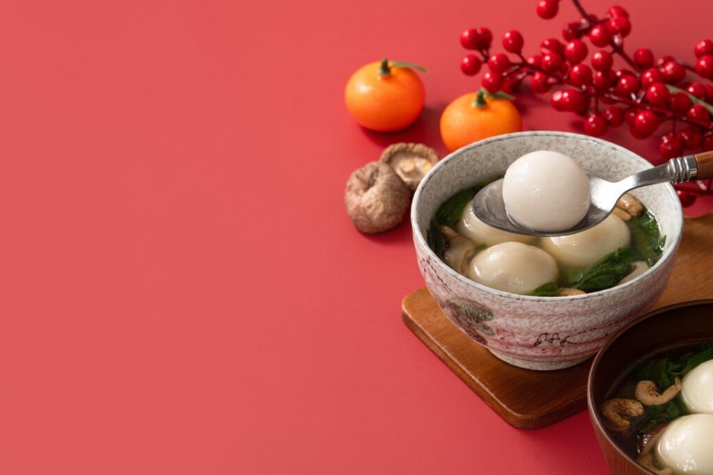 A bowl of Tangyuan soup, traditional Chinese New Year glutinous rice balls, served with clementines and red berries on a bright red background.