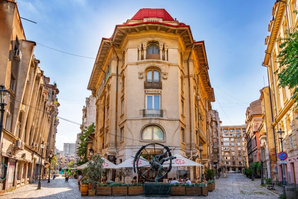 A beautiful historic building with a red roof located at a street corner in the Lipscani district of Bucharest, featuring an outdoor cafe and a modern sculpture in the foreground.