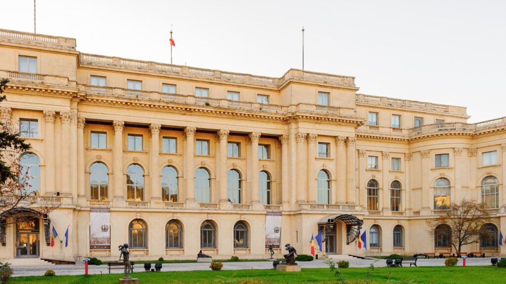 The grand facade of the National Museum of Art of Romania in Bucharest, featuring classical columns and statues on a green lawn under a clear sky.