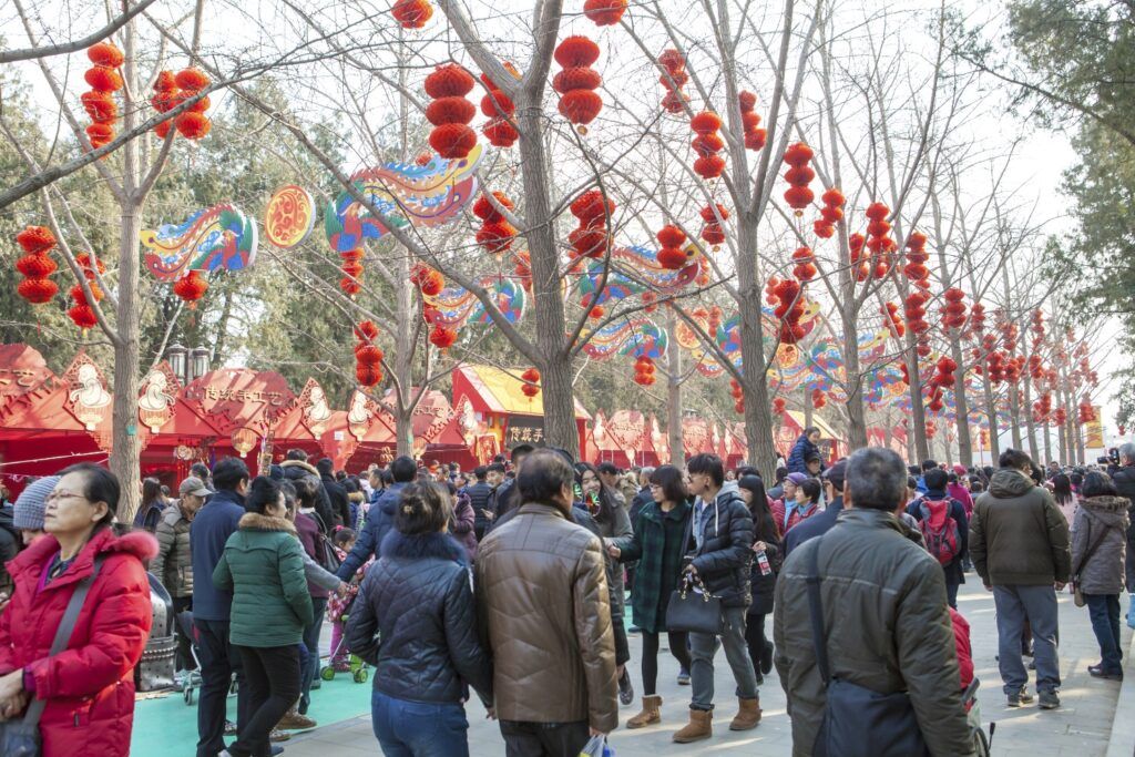 A crowd of people celebrates the Chinese New Year in Beijing, walking under trees decorated with traditional red lanterns.