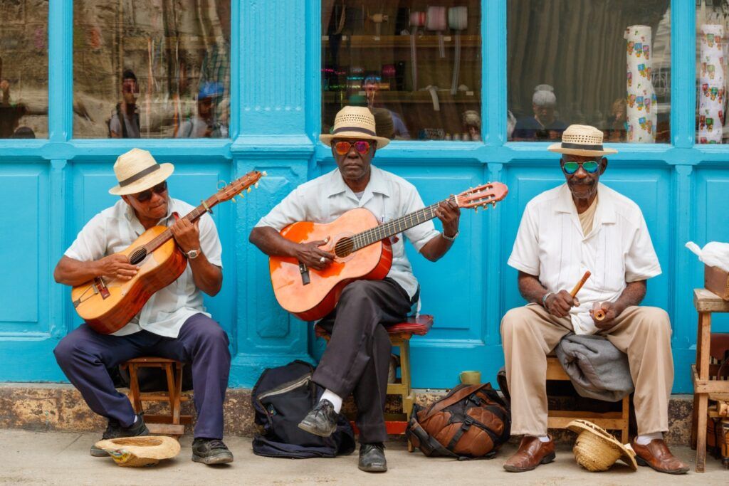 Drei Straßenmusiker sitzen auf Hockern vor einem strahlend blauen Gebäude in Havanna und spielen Gitarre und Percussion, während sie Strohhüte und Sonnenbrillen tragen.