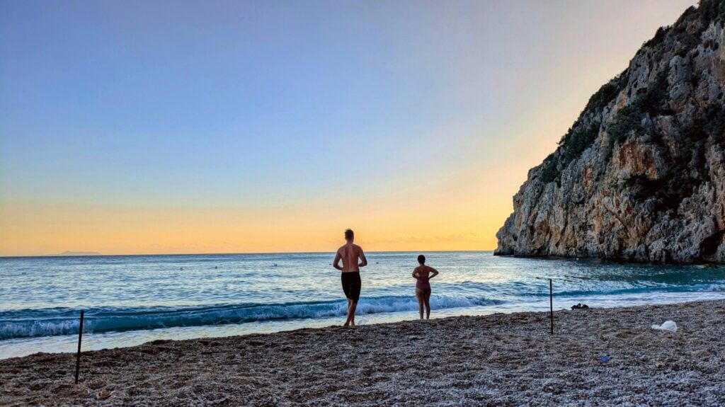 Two people standing on the pebbly shore of Dhermi Beach in Albania, looking out at the calm sea during a golden sunset.